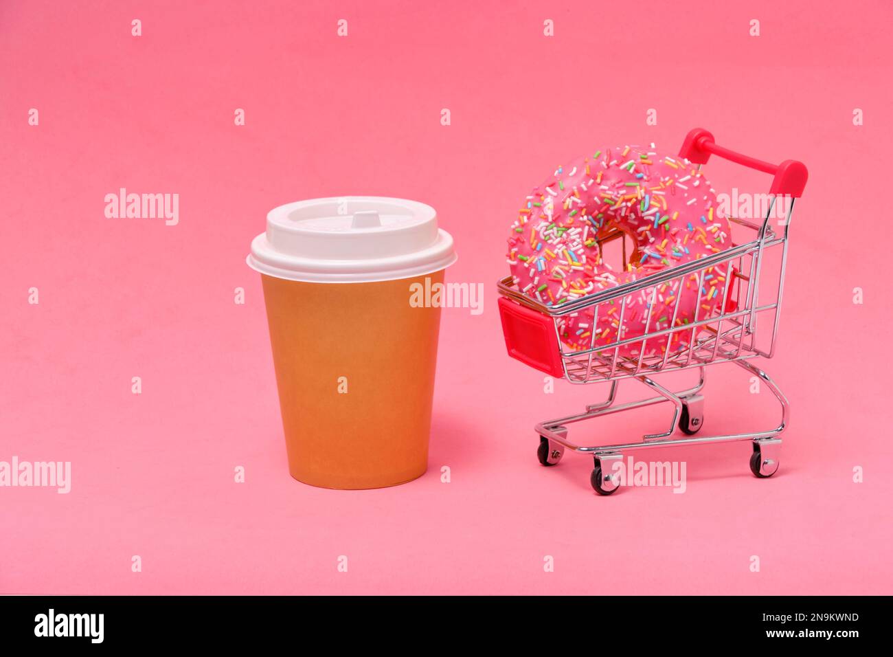 Ciambella rosa nel carrello della spesa e tazza da caffè usa e getta su sfondo rosa Foto Stock
