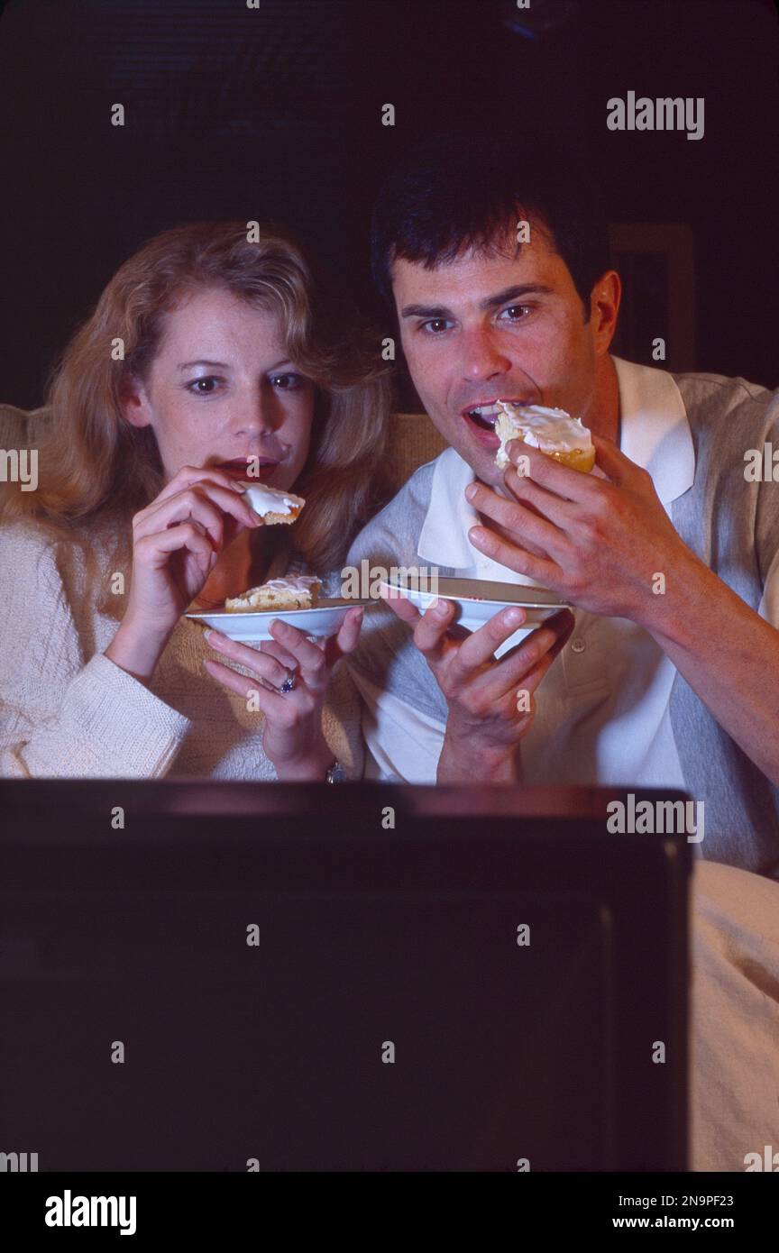 Primo piano della coppia più giovane che ripieno le loro facce con cibo spazzatura mentre guardano la televisione Foto Stock