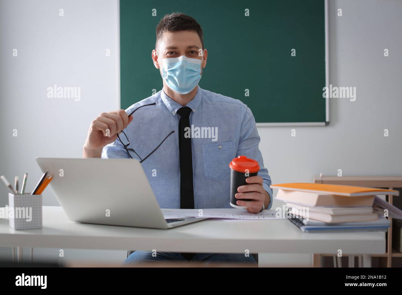 Insegnante con maschera protettiva e computer portatile seduto alla scrivania in aula. Riapertura dopo la quarantena di Covid-19 Foto Stock