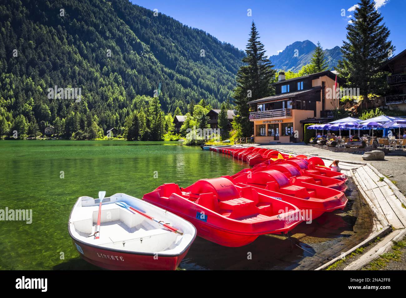 Barche rosse allineate al lago Champex con un caffè all'aperto sotto il cielo blu; Champex, Vallese, Svizzera Foto Stock