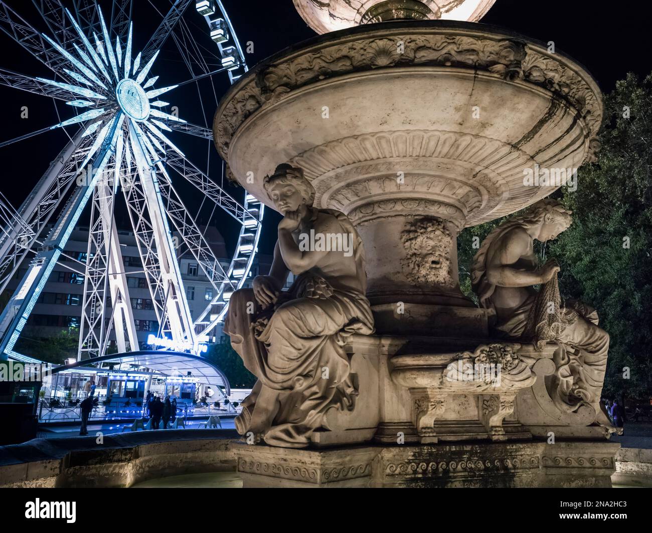 Una ruota e un edificio illuminati dei traghetti con una statua in pietra scolpita in primo piano di notte; Budapest, Budapest, Ungheria Foto Stock