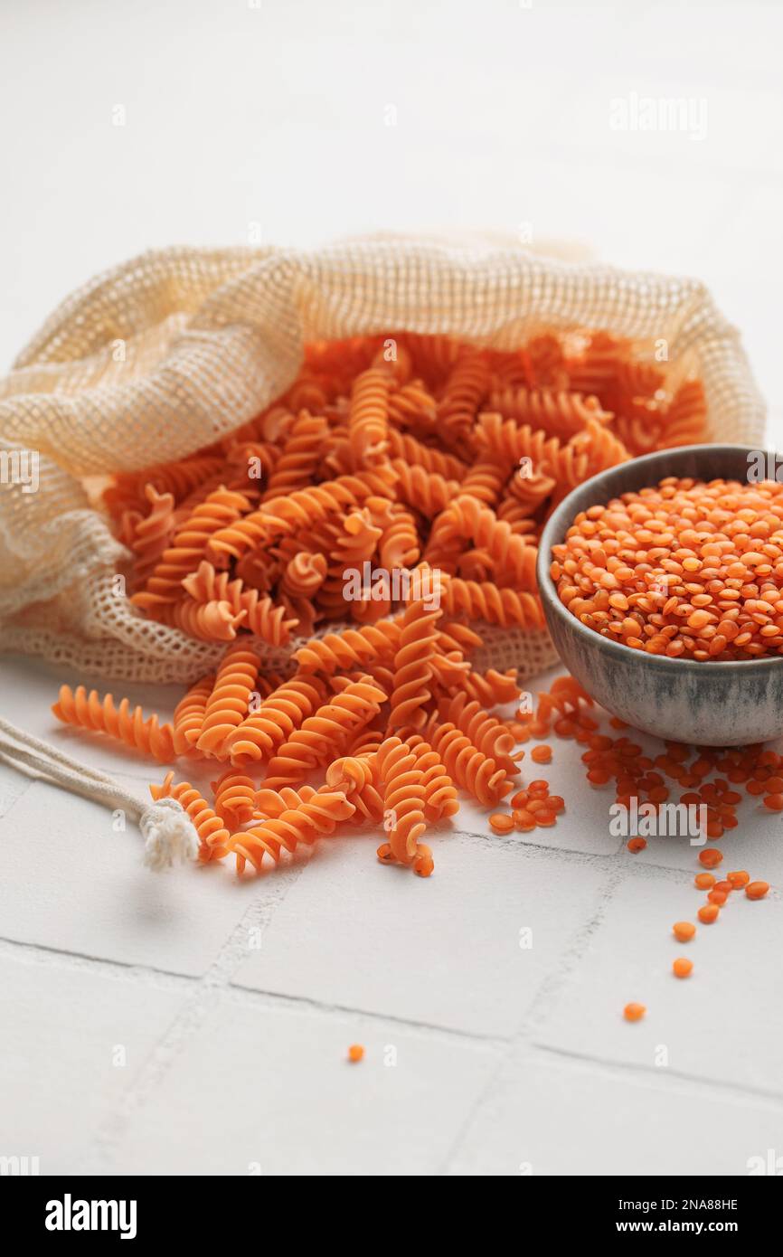 Lenticchie rosse fusilli su fondo piastrellato bianco. Sacchetto con pasta cruda e ciotola con lenticchie rosse. Pasta senza glutine. Foto Stock