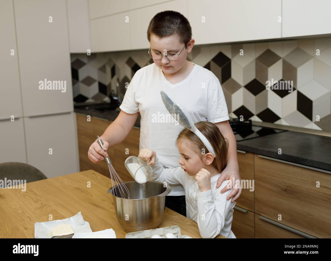 Fratelli cucinano, preparano e mescolano la pasta, facendo sorpresa, aiutando i genitori. Fratello e sorella in orecchie conigliate in piedi insieme al tavolo da cucina dentro Foto Stock