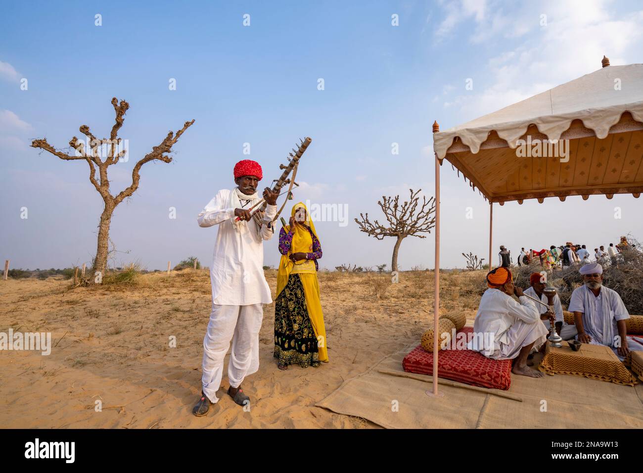 Rajput uomo che suona ravanahatha e cantante donna che si esibisce al festival con anziani del villaggio che fumano un narghilè nel deserto del Thar nel Rajasthan, India Foto Stock