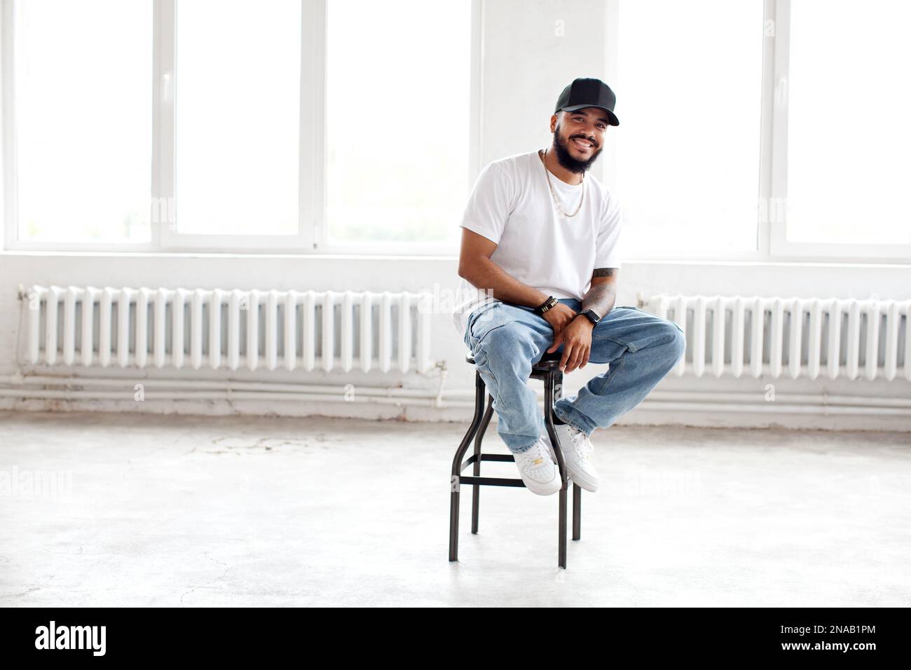 Autentico Ritratto felice uomo portatore seduto in bianco sfondo loft, sorridente, indossando casual T shirt bianca, berretto da baseball e jeans, lifestyle Foto Stock