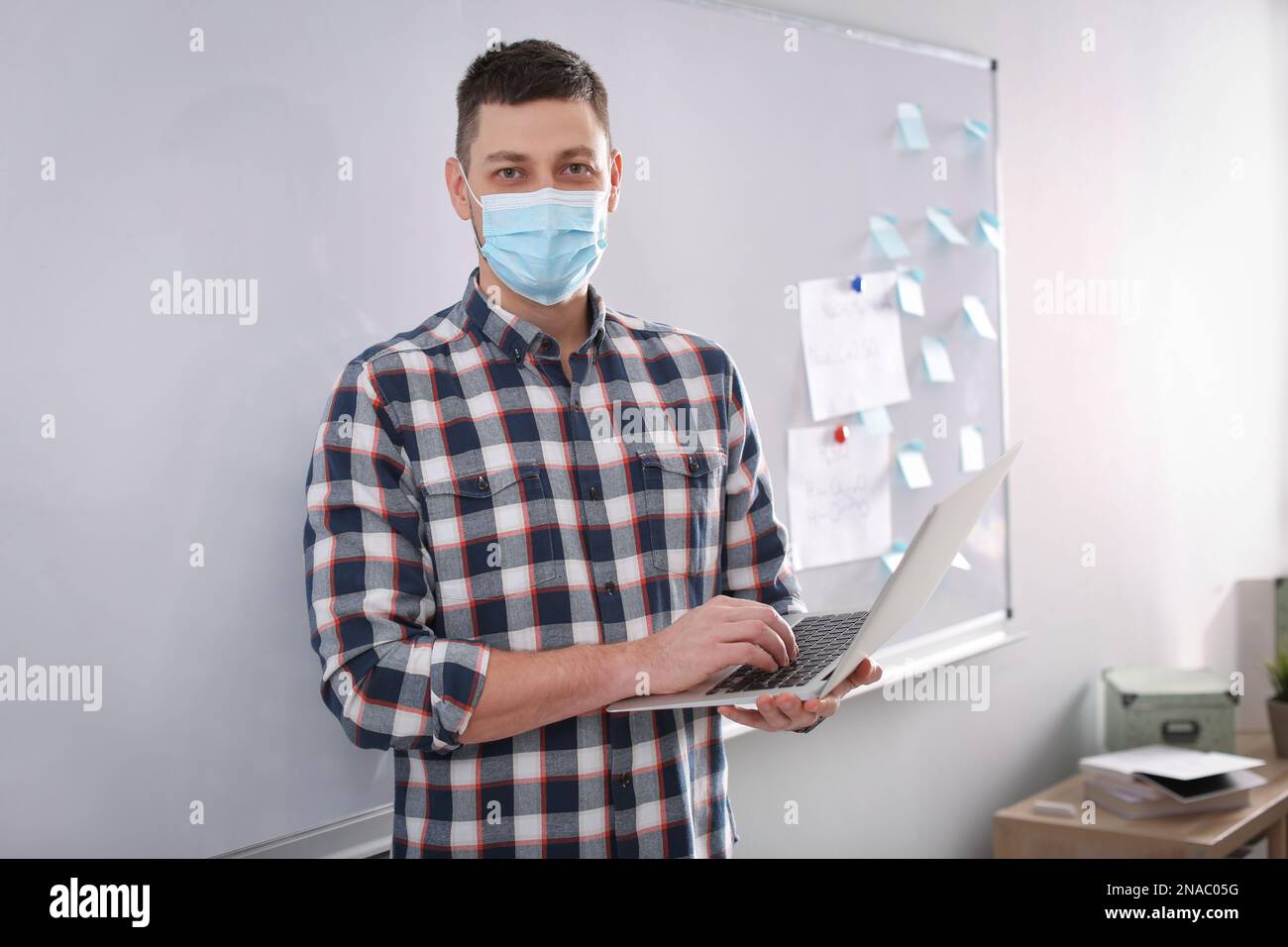 Insegnante con maschera protettiva e computer portatile vicino al bordo in aula. Riapertura dopo la quarantena di Covid-19 Foto Stock