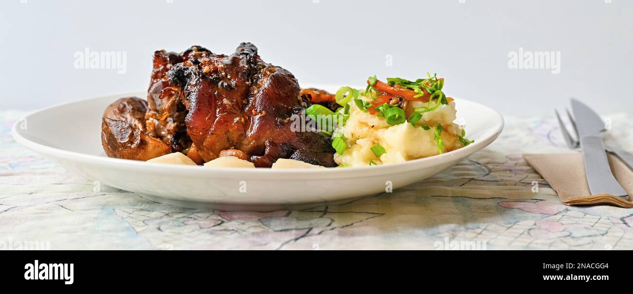 Ginocchio succoso di maiale al forno con purè di patate e verdure sul piatto, posate sul tavolo, primo piano. Foto Stock