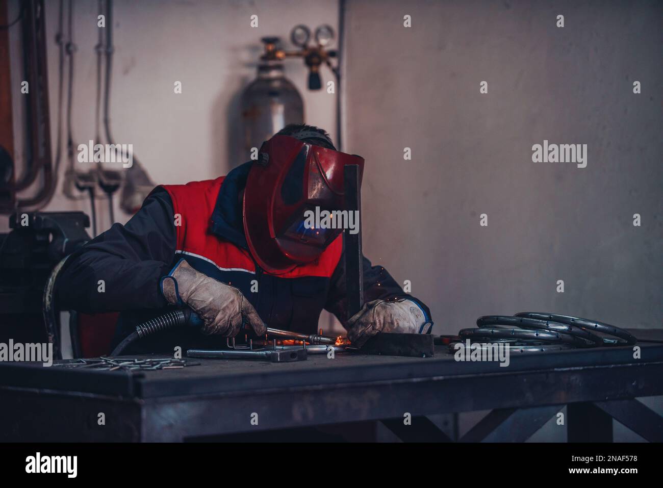 Saldatrice professionale in uniforme protettiva e maschera saldante tubo metallico sul tavolo industriale con altri lavoratori dietro nell'officina industriale. Foto Stock