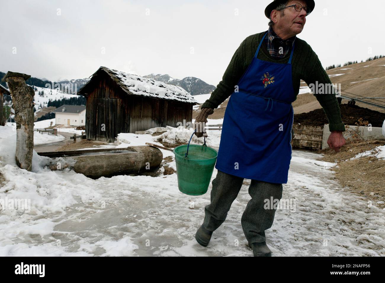 L'uomo ladino raccoglie un secchio d'acqua da una fossa di bestiame e attraversa con attenzione un foglio di ghiaccio. La vita e' dura in villaggi rurali e isolati, lik... Foto Stock