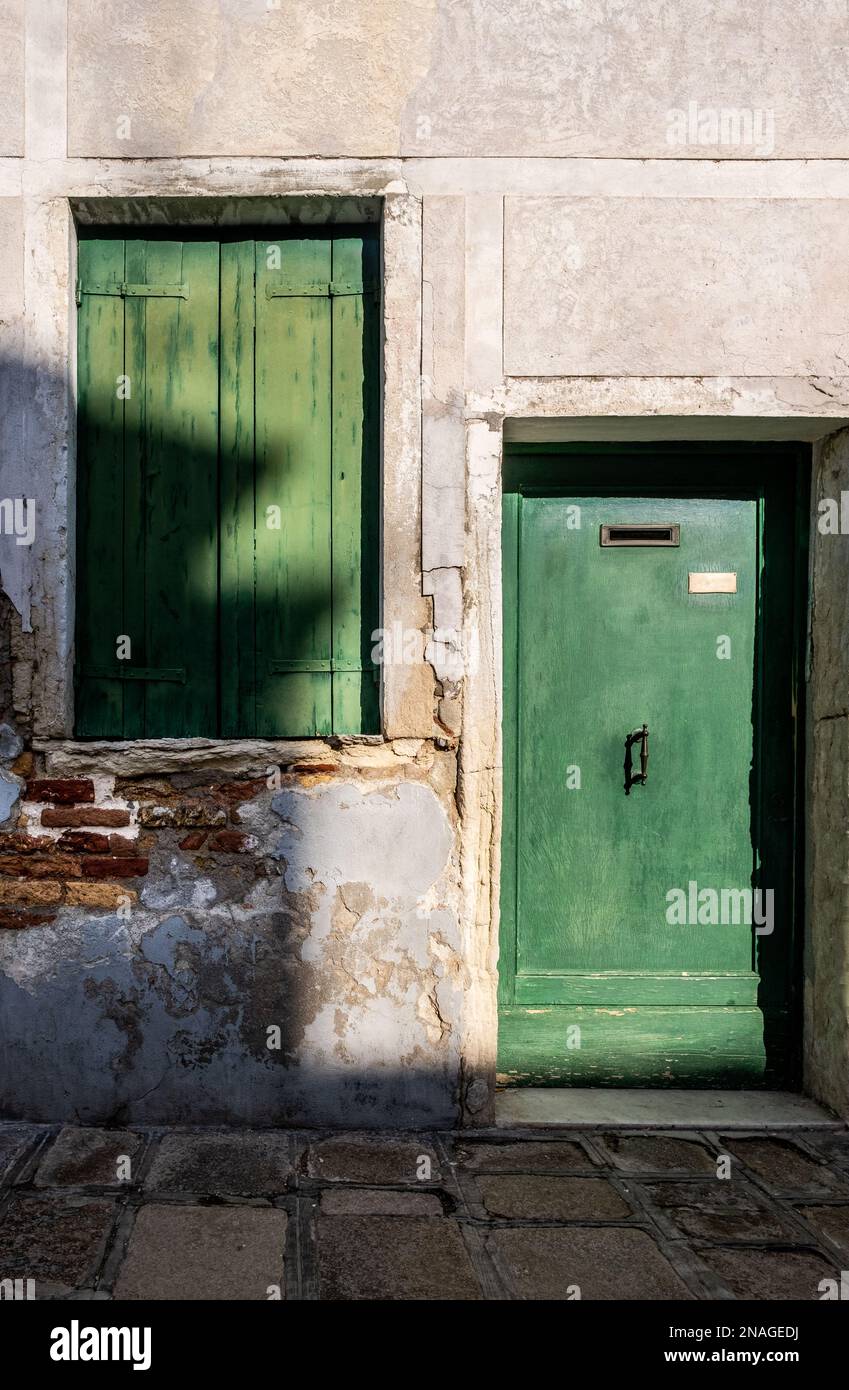 Forte luce del sole che proietta ombre attraverso una finestra grezza chiusa e una porta verde in una delle case di Burano, Venezia, Italia Foto Stock