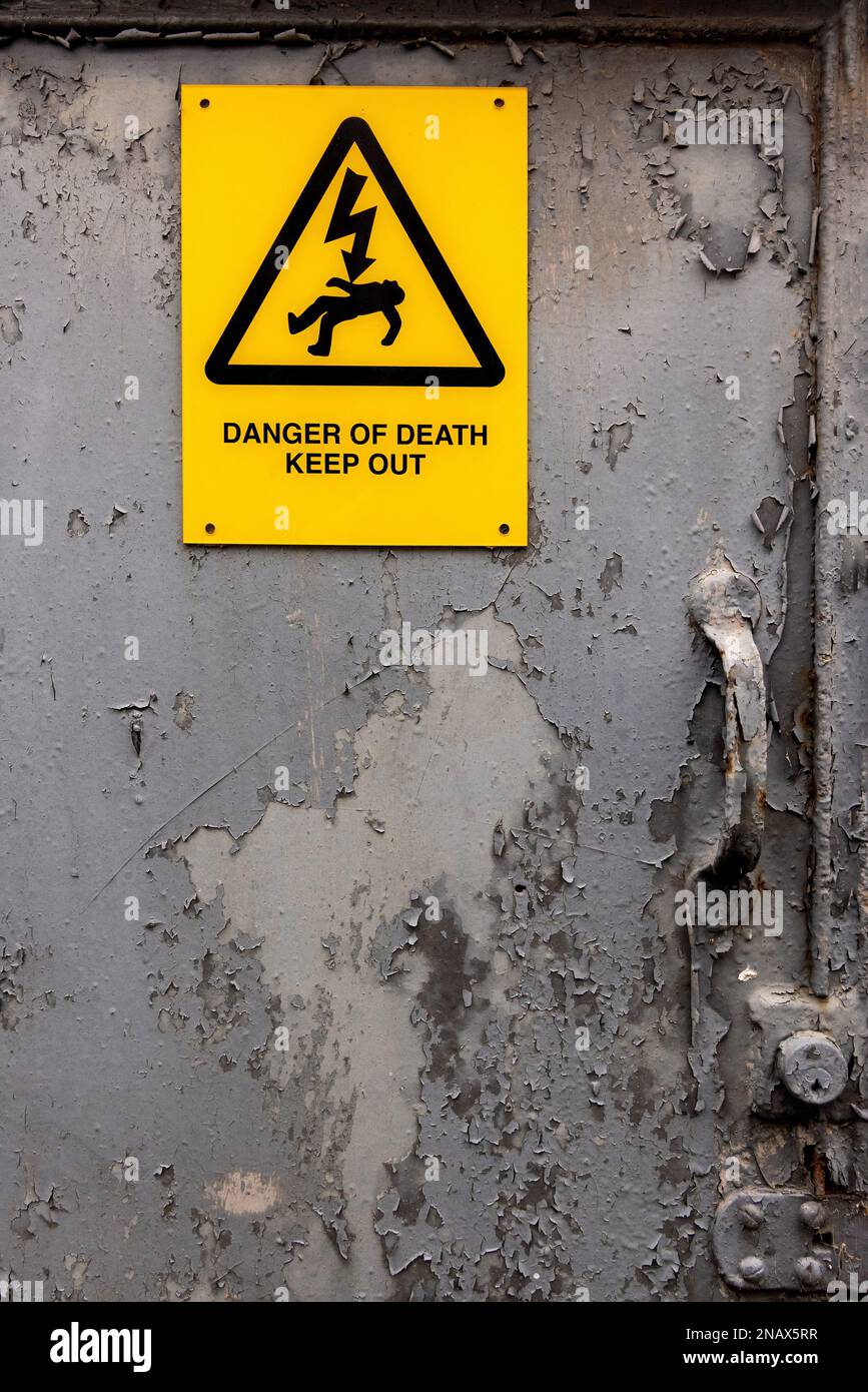 Avviso di pericolo elettrico con la scritta "Pericolo di morte – Vietato l'ingresso" su una vecchia porta metallica corrosa con vernice grigia scrostata. Foto Stock