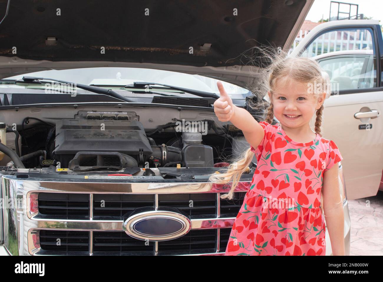 Felice bambina caucasica in piedi vicino al cofano aperto della macchina, mostrando un buon segno. gesto della mano Foto Stock