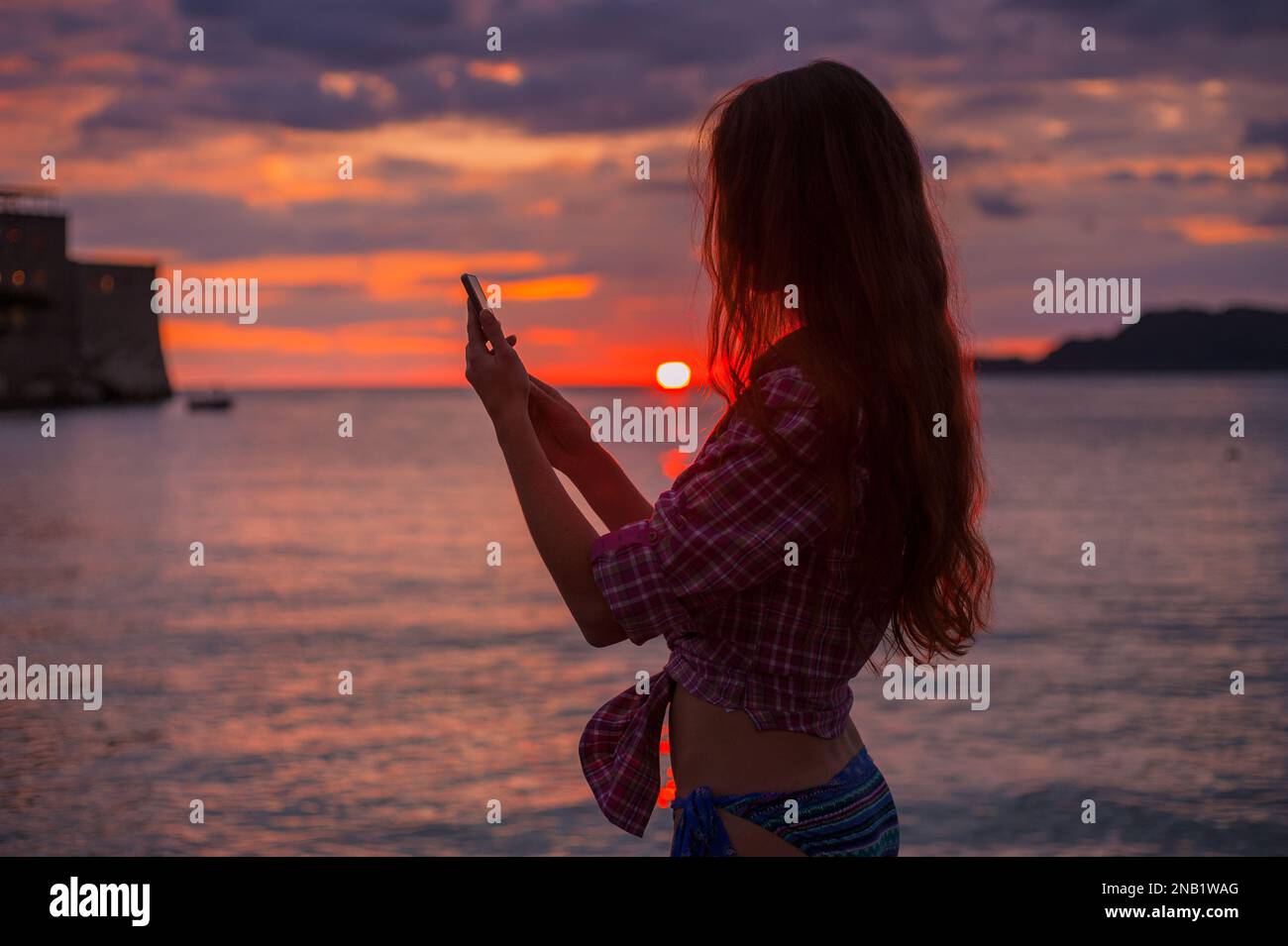 Vista laterale silhouette una giovane donna rossa che usa lo smartphone sulla spiaggia durante l'idilliaco e colorato tramonto sul mare Adriatico, foto scattata a Budva Montenegro Foto Stock