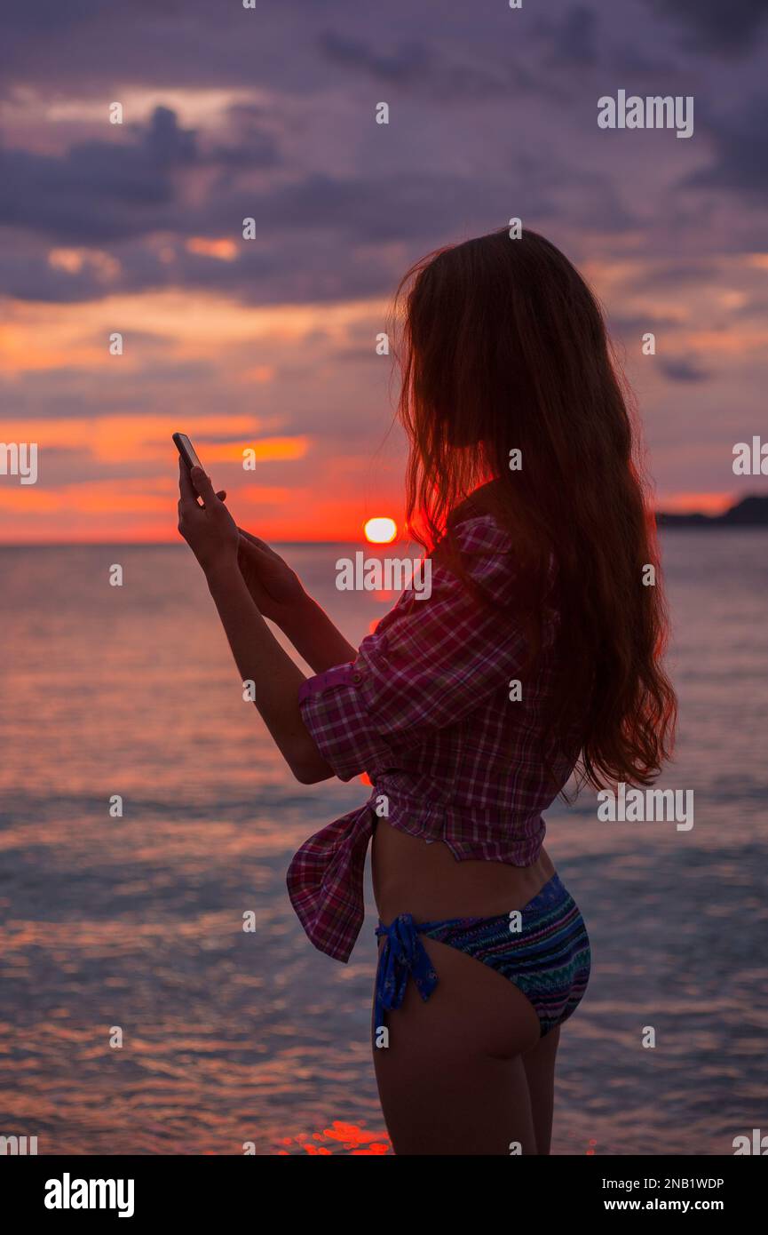 Vista laterale silhouette una giovane donna rossa che usa lo smartphone sulla spiaggia durante l'idilliaco e colorato tramonto sul mare Adriatico, foto scattata a Budva Montenegro Foto Stock