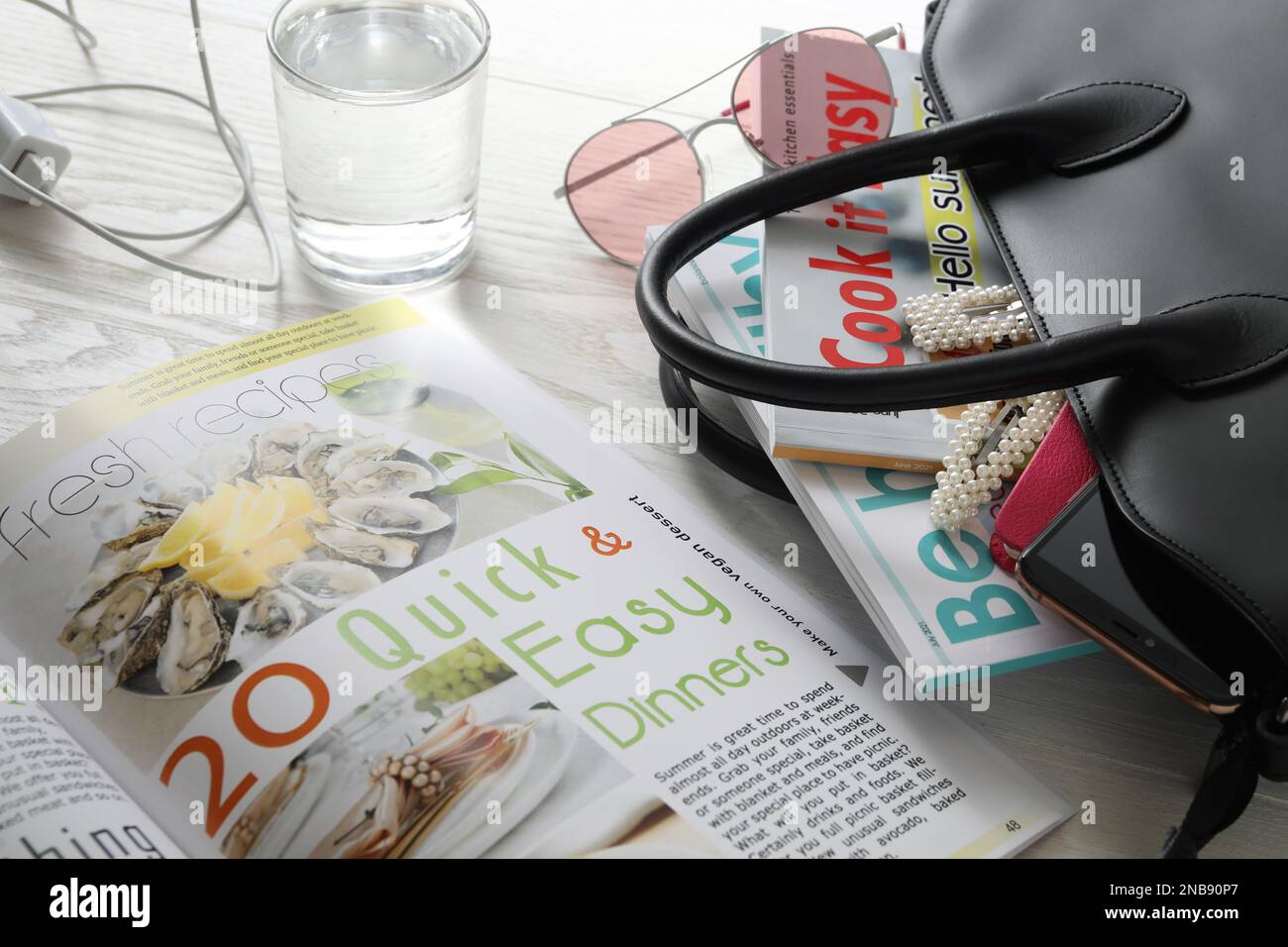 Borsa con diversi caricatori, accessori e bicchiere d'acqua su tavolo di legno bianco, primo piano Foto Stock