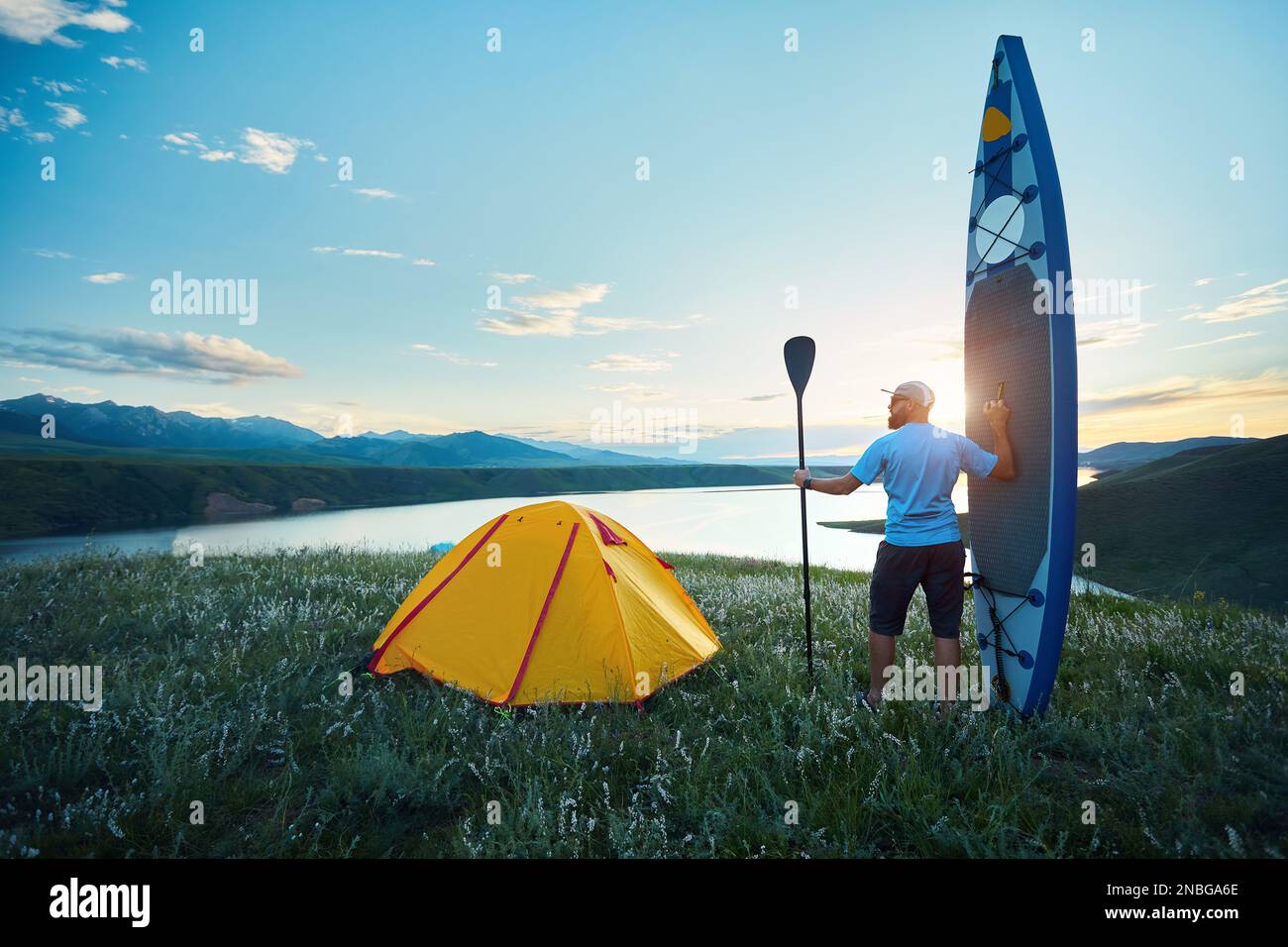 Uomo con pagaia e SUP bordo vicino tenda gialla campeggio lago al tramonto in Kazakistan. Stand up paddle board attività ricreative all'aperto in natura. Foto Stock