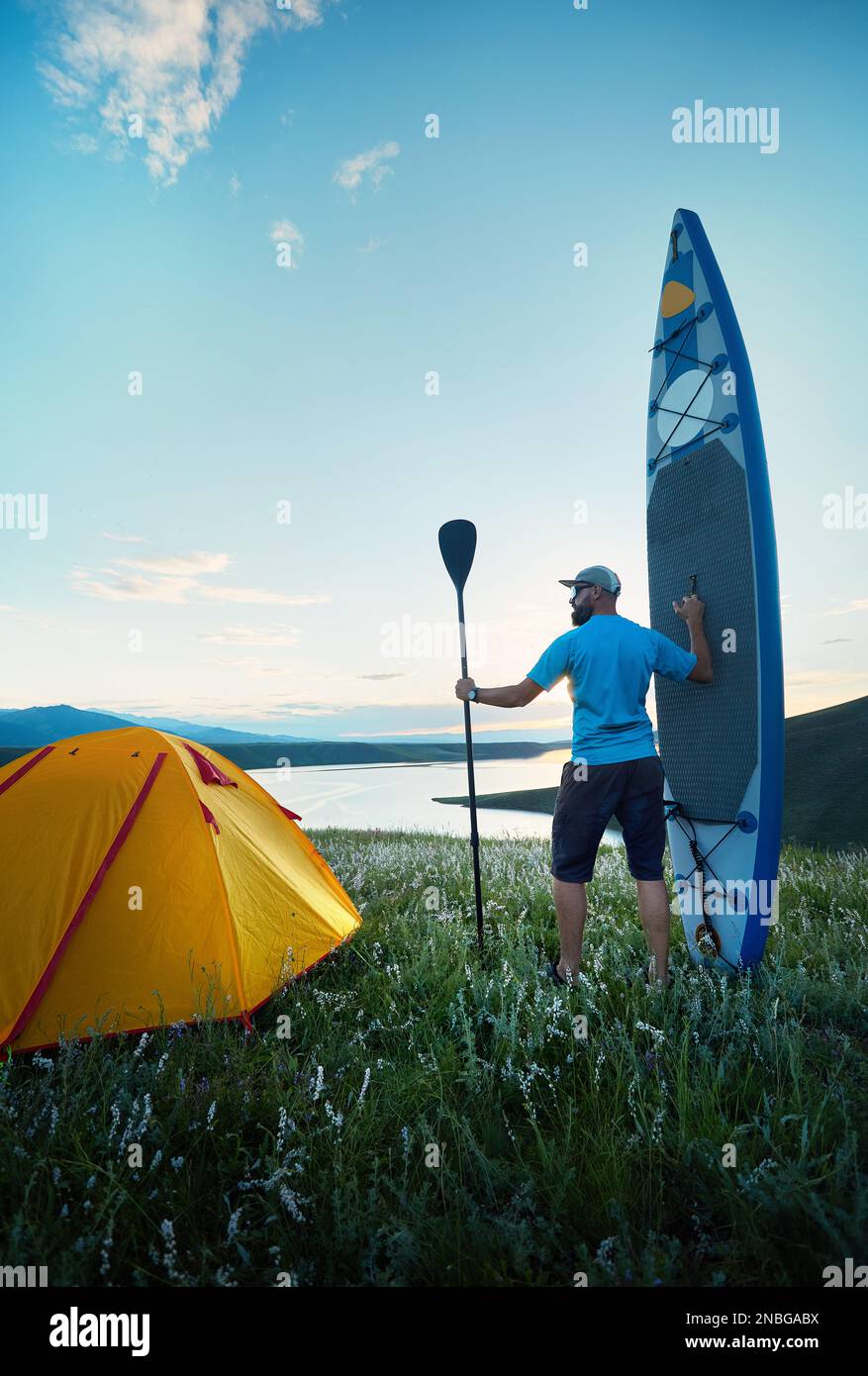 Uomo con pagaia e SUP bordo vicino tenda gialla campeggio lago al tramonto in Kazakistan. Stand up paddle board attività ricreative all'aperto in natura. Foto Stock