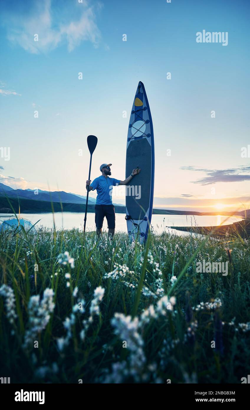Uomo con pagaia e SUP bordo vicino tenda blu campeggio lago al tramonto in Kazakistan. Stand up paddle board attività ricreative all'aperto in natura. Foto Stock