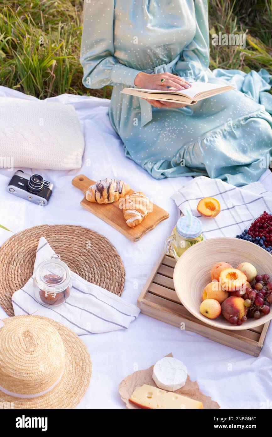 Foto ritagliata della donna in abito lungo con capelli corti seduti su una coperta bianca con frutta e dolci e leggendo il libro. Concetto di avere picni Foto Stock