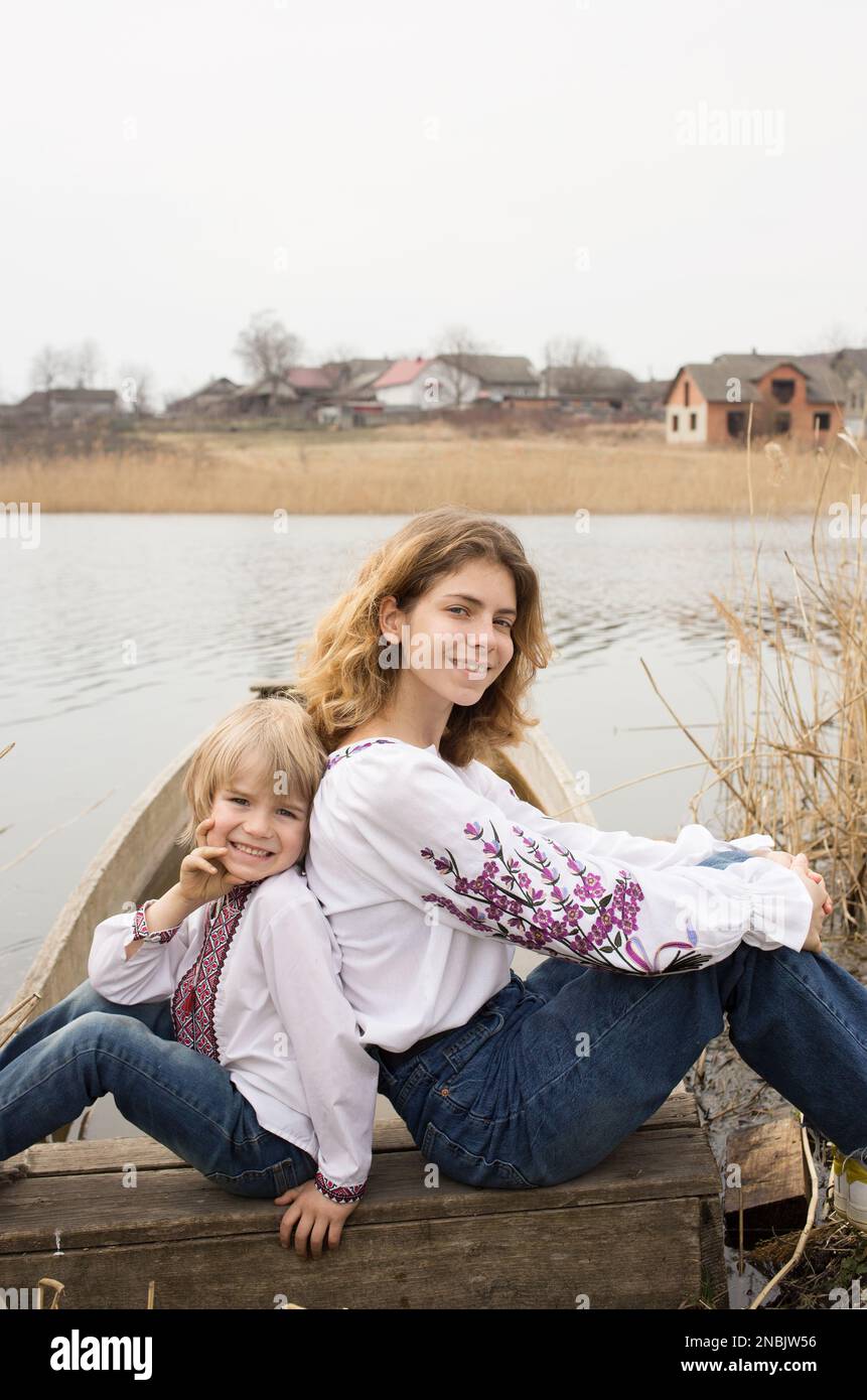 felice ragazzino e giovane bella donna in abiti ricamati nazionali siedono accanto l'uno all'altro sulla riva del lago. Famiglia, rifugiati, unità, sostegno, Foto Stock