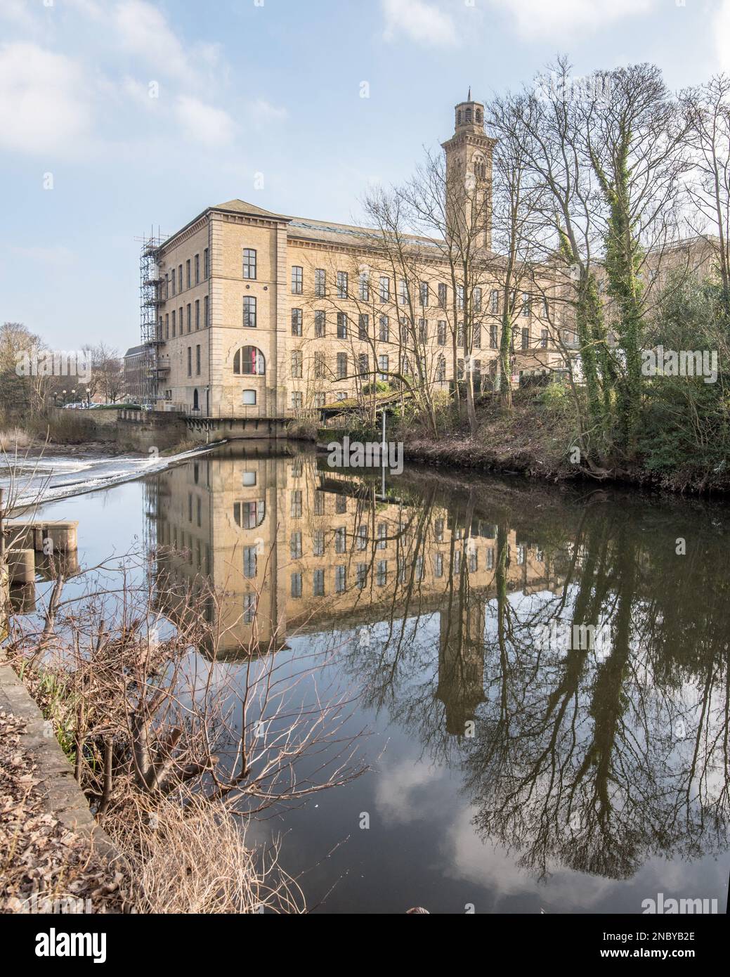 Riflesso dell'edificio NHS 'New Mill' nella superficie dell'acqua sopra lo stramazzo e sotto il Victoria Bridge, Saltaire, West yorkshire. Foto Stock