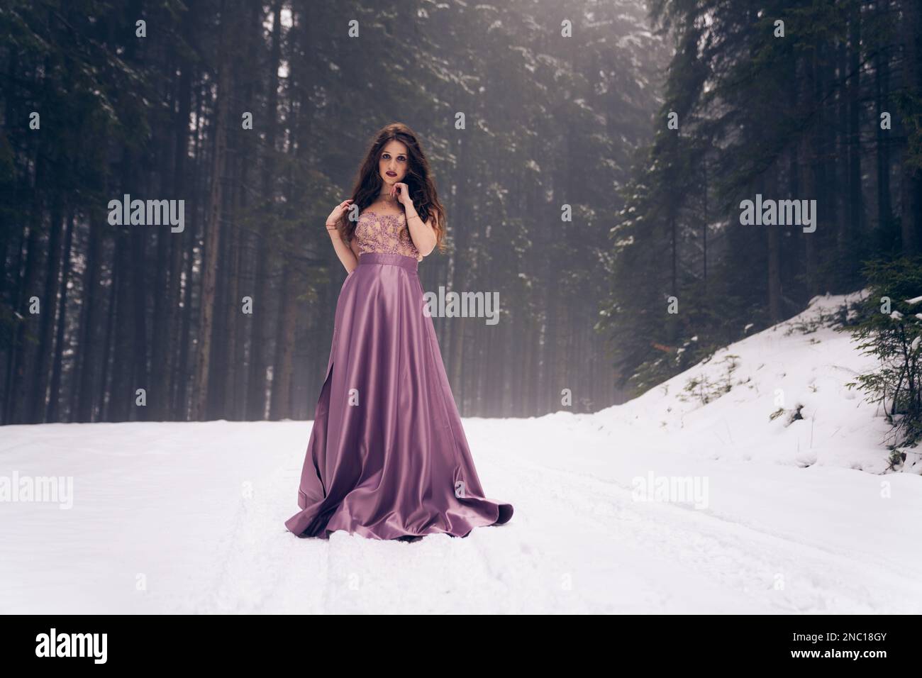 Una donna in un abito di raso viola contro il paesaggio invernale, foresta innevata sentiero Foto Stock