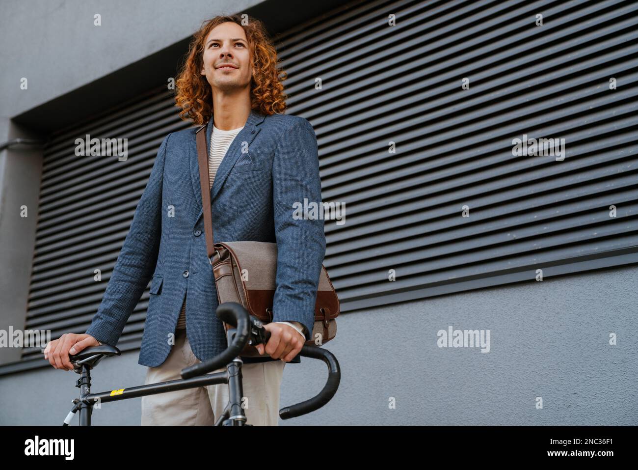 Giovane zenzero uomo a capelli lunghi in piedi con bicicletta vicino a parete grigia all'aperto Foto Stock