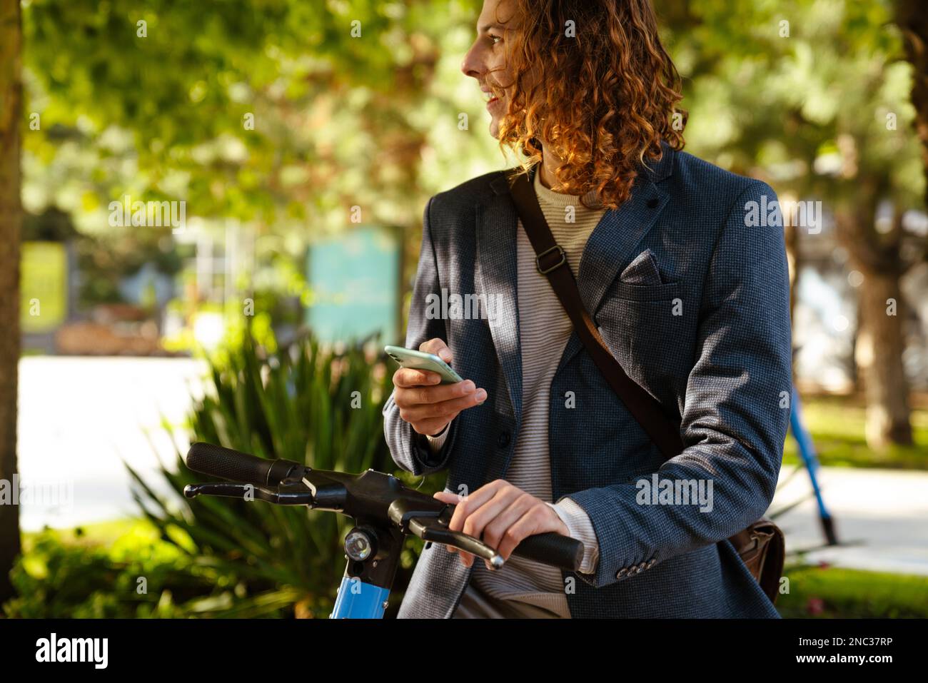 Zenzero uomo europeo utilizzando il cellulare mentre si è in piedi con scooter elettrico al parco Foto Stock