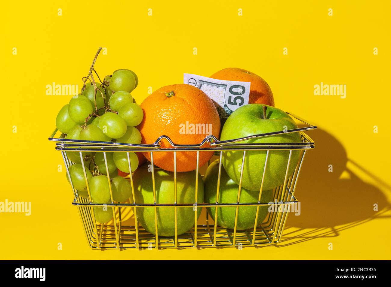 cestino supermercato carrello con frutta e dollari, frutta per cibo e denaro, carta rotonda multicolore, si trovano in un cestino sulla superficie del giallo Foto Stock