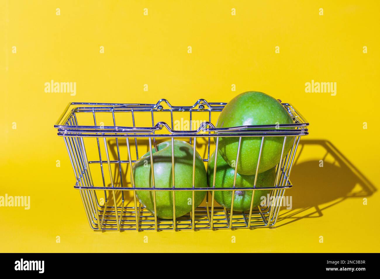 carrello con mele, frutta per il cibo, carrello supermercato, diversi oggetti, verde rotondo, griglia di metallo, si trova sulla superficie dello sfondo giallo. Foto Stock