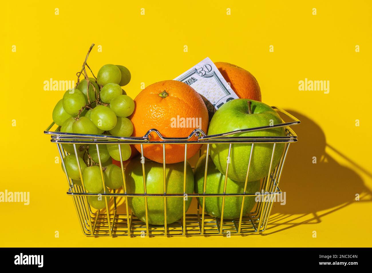 cestino supermercato carrello con frutta e dollari, frutta per cibo e denaro, carta rotonda multicolore, si trovano in un cestino sulla superficie del giallo Foto Stock