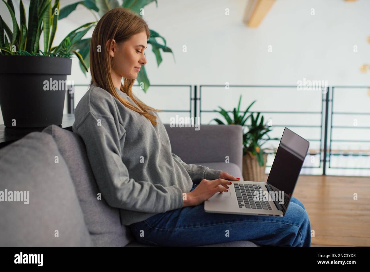 Vista laterale in felice bionda giovane donna alla ricerca di un moderno schermo notebook seduto su un comodo divano a casa, mani al lavoro con tecnologia digitale Foto Stock