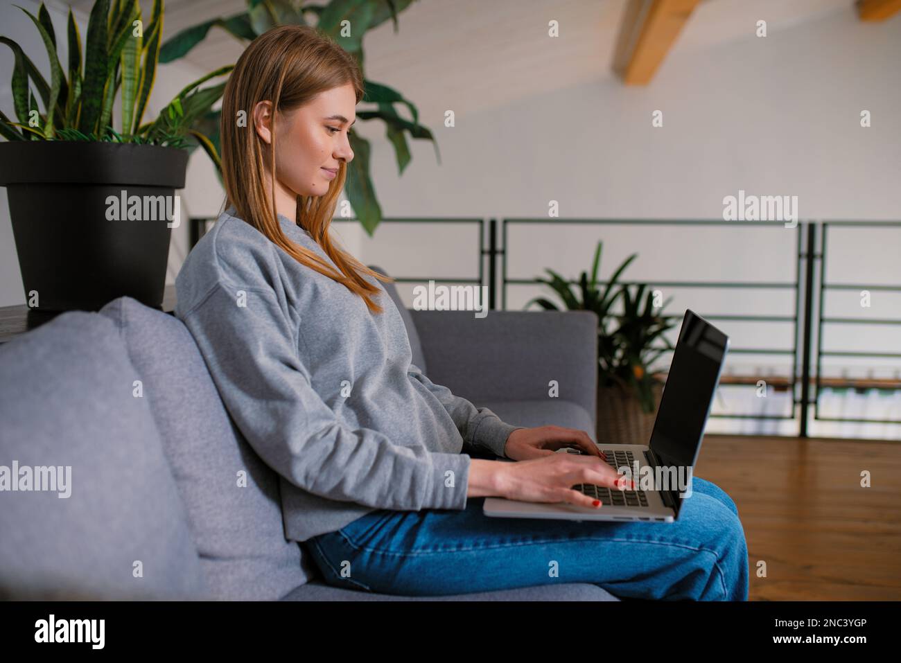Vista laterale in felice bionda giovane donna alla ricerca di un moderno schermo notebook seduto su un comodo divano a casa, mani al lavoro con tecnologia digitale Foto Stock