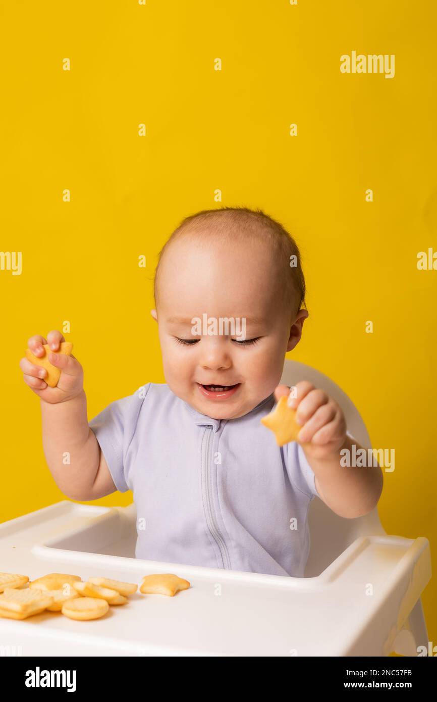 un bambino cute sta sedendosi nella sedia del bambino che mangia i biscotti. Sfondo giallo. Pappe per bambini, spazio per il testo. Foto di alta qualità Foto Stock
