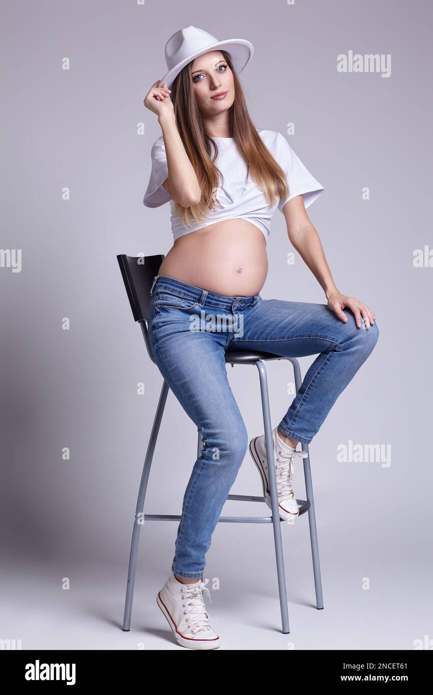Giovane donna incinta in t-shirt bianca, e jeans sedersi su sgabello bar. Femmina con pancia esposta. 5th mese di gravidanza. Foto Stock