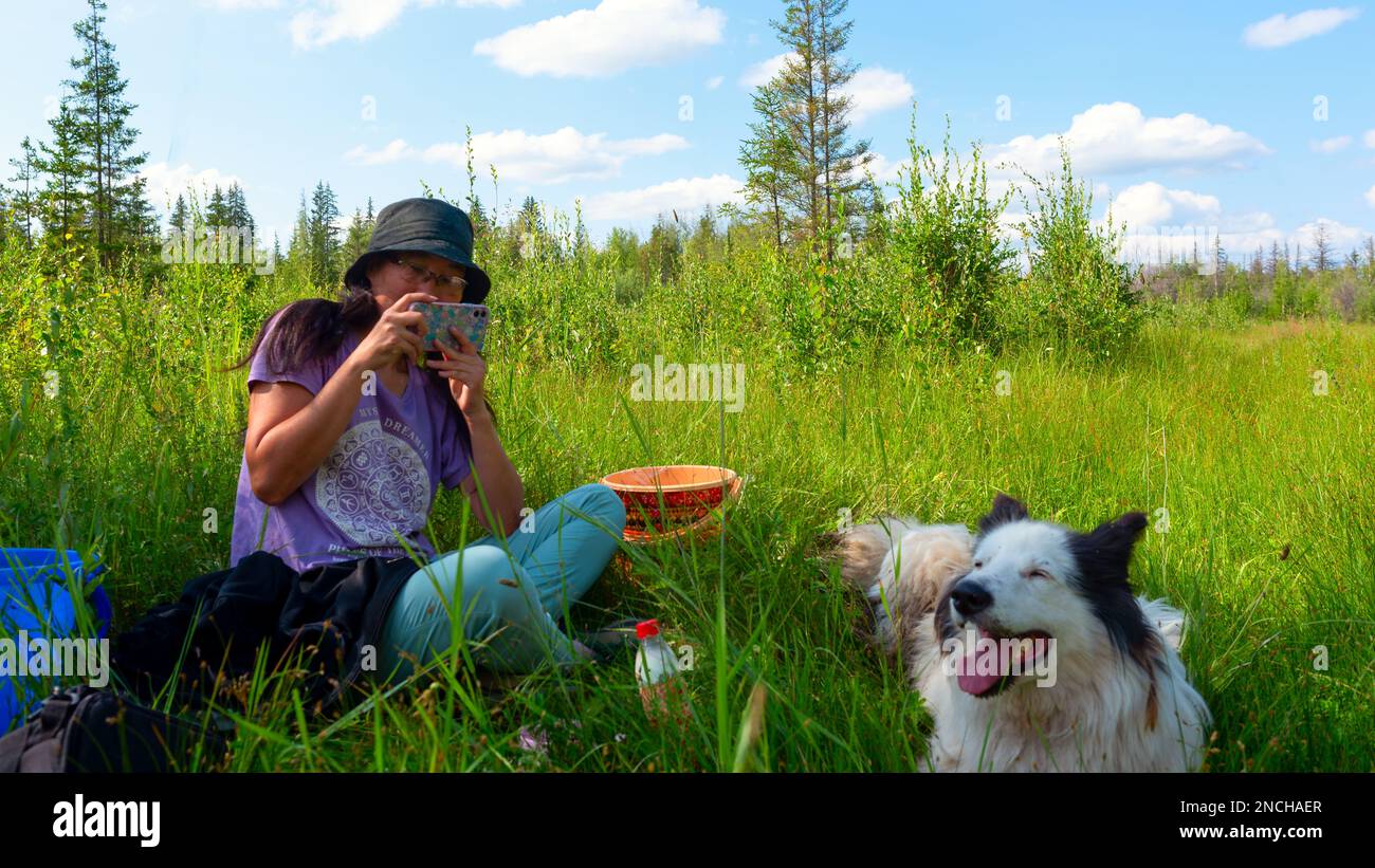 Una ragazza in un cappello seduta sull'erba verde scatta una foto al telefono con un cane bianco gioioso nella foresta durante il giorno. Foto Stock