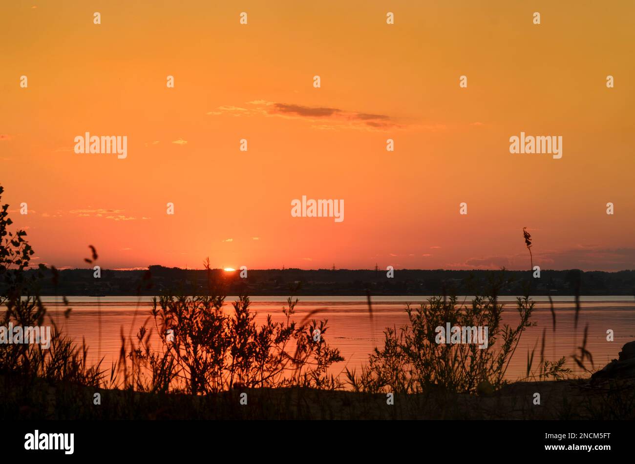 Parte del luminoso disco solare sul tramonto arancione all'orizzonte nel cielo sopra il fiume d'erba sopra le case del villaggio in Russia. Foto Stock