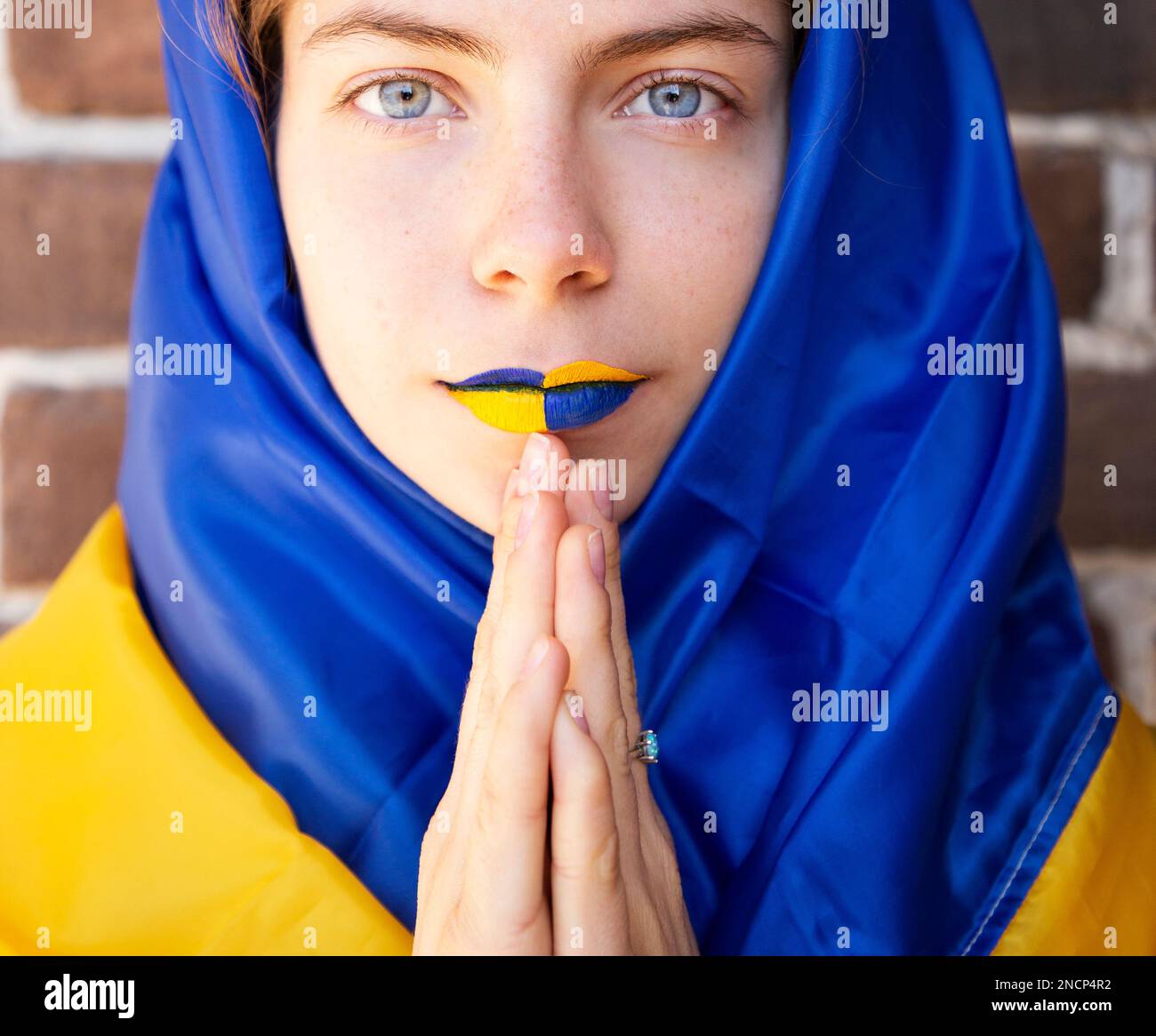 Bel volto di una giovane donna Ucraina con labbra dipinte giallo-blu e mani piegate in preghiera. Uno sguardo penetrante. fermare la guerra. Speranza e fede in v Foto Stock