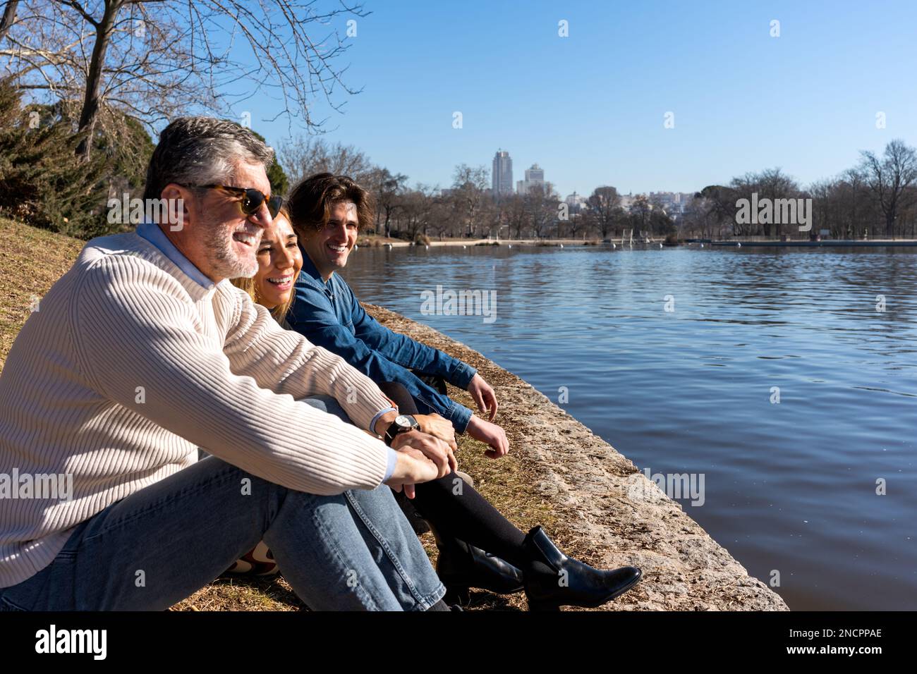 Vista laterale corpo completo di positivo uomo maturo grigio con capelli in occhiali da sole seduti con moglie e figlio vicino al fiume, mentre si passa il fine settimana insieme Foto Stock