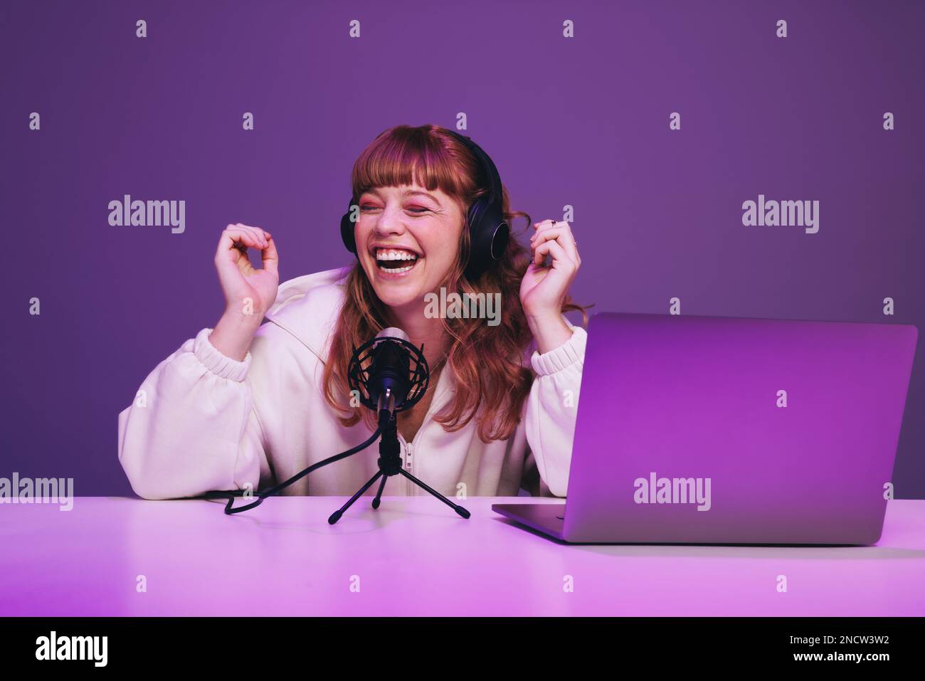 Podcaster femminile ridere mentre ospita una trasmissione dal vivo in uno studio. Felice giovane donna che registra uno spettacolo audio in luce viola al neon. Donna che crea c Foto Stock