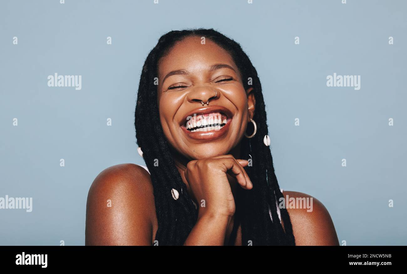 Donna africana con piercing viso sorridente allegro in uno studio. Felice giovane donna che indossa Dreadlocks e gioielli piercing contro un backgroun blu Foto Stock