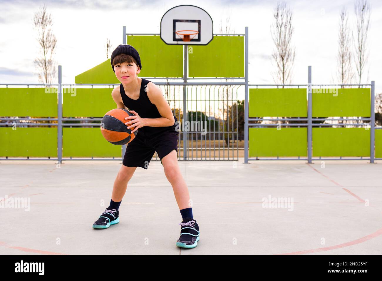 Corpo pieno di giovane bambino sportivo in abbigliamento sportivo e cappello dribbling palla mentre si pratica il basket sul campo contro cerchio durante l'allenamento Foto Stock