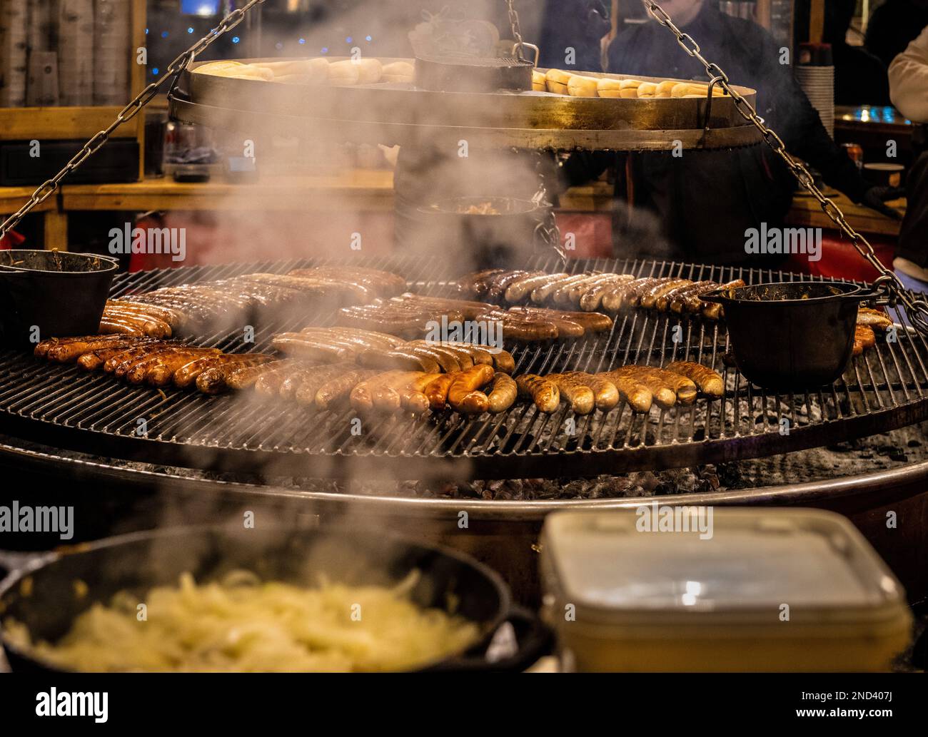Salsicce che cucinano su una griglia circolare sospesa sopra un barbecue, con vapore da una padella di cipolle in primo piano. Foto Stock
