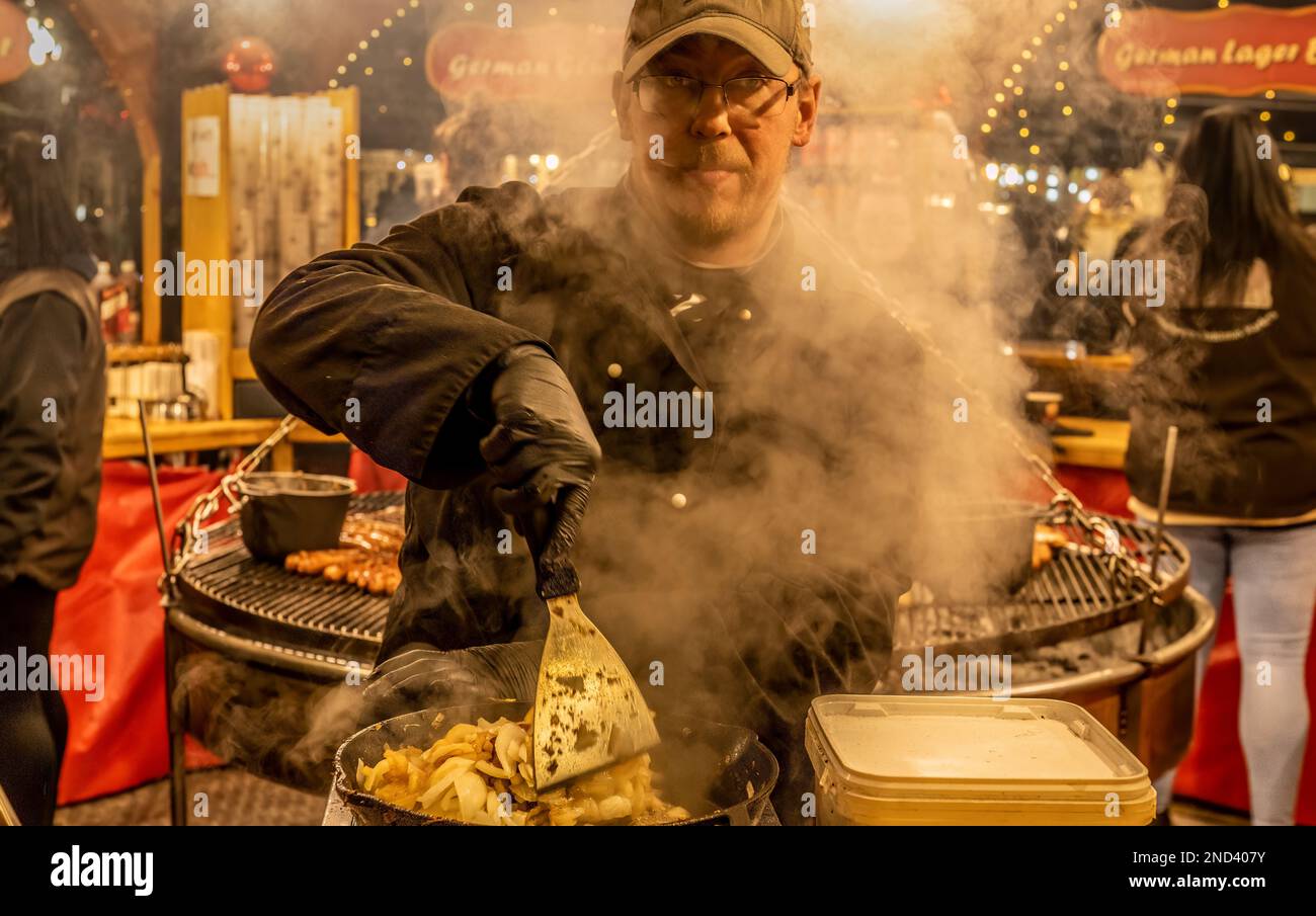 Un uomo friggerà un grande recipiente di cipolle fumanti, con un grande grill rotondo sospeso e barbecue dietro di lui. Mercatino di Natale di Manchester. REGNO UNITO Foto Stock