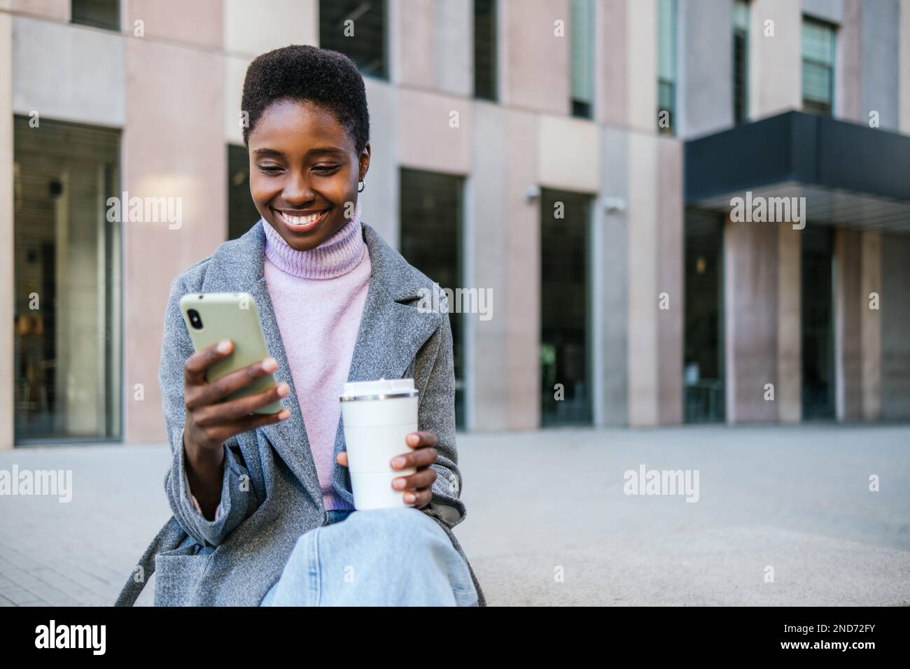 Donna afro-americana ottimista in outerwear elegante con tazza di caffè eco-friendly scorrere le notizie sullo smartphone mentre si siede sulla panca in città strada Foto Stock