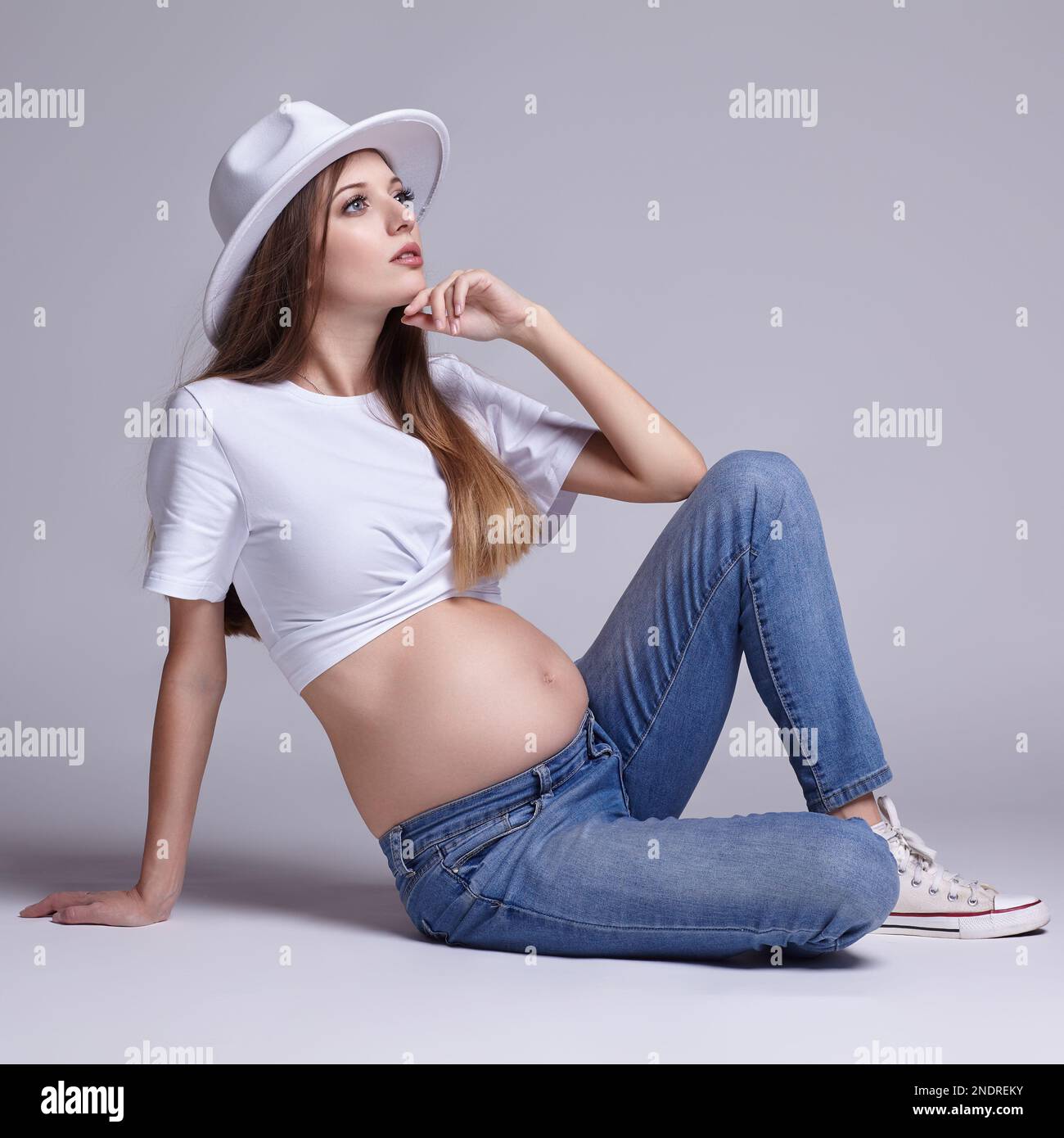 Giovane donna incinta in t-shirt bianca, cappello e jeans seduti sul pavimento sullo sfondo. Femmina con pancia esposta. 5th mese di gravidanza. Foto Stock