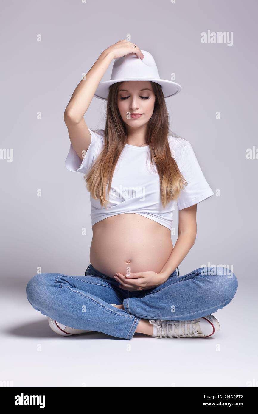 Giovane donna incinta in t-shirt bianca, cappello e jeans seduti sul pavimento sullo sfondo. Femmina con pancia esposta. 5th mese di gravidanza. Foto Stock