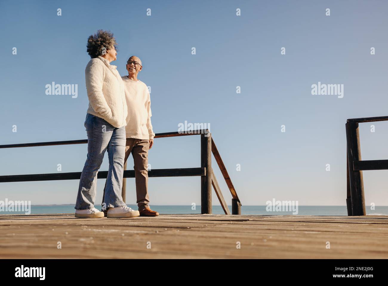 Vista ad angolo basso di una coppia matura sorridente mentre si fa una passeggiata lungo un ponte pedonale sulla spiaggia. Romantica coppia anziana godendo gli spendi Foto Stock