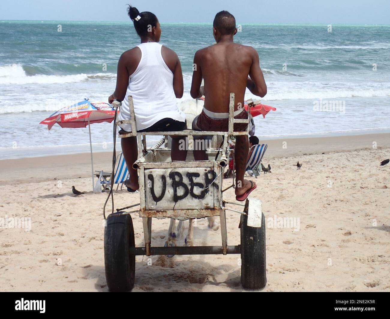 Una coppia in bicicletta un carrello da spiaggia con un cartello fatto in casa che dice Uber su di esso, sulla spiaggia di Salvador, Brasile. Foto Stock