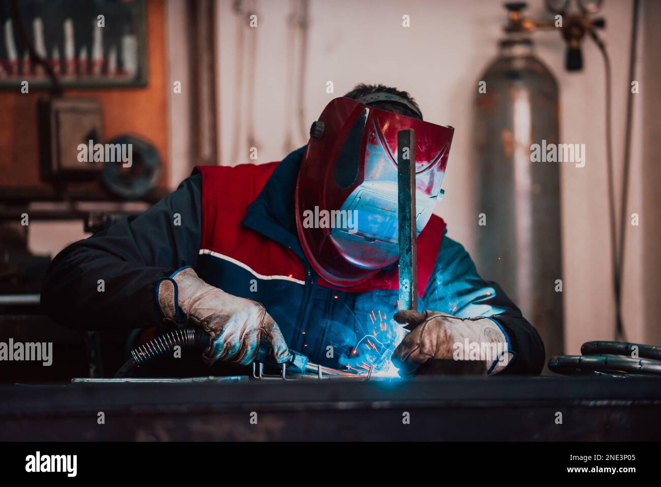 Saldatrice professionale in uniforme protettiva e maschera saldante tubo metallico sul tavolo industriale con altri lavoratori dietro nell'officina industriale. Foto Stock